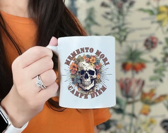 May include: White ceramic mug with a black and white skull design surrounded by colorful flowers. The text "Memento Mori Carpe Diem" is printed on the mug.