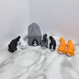 Op de afbeelding: Een set van zeven kattenfiguren, waaronder een grijs kattenhuis, een zwart-witte kat, een witte kat, een grijze kat en drie oranje katten. De figuren staan allemaal in een speelse pose.