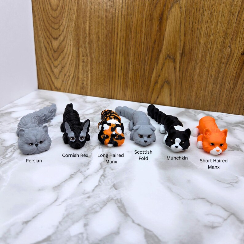 Op de afbeelding: Een set van zeven 3D-geprinte kattenfiguren, elk representatief voor een ander ras. De katten zijn in verschillende poses, waaronder liggend, zittend en staand. De figuren zijn geschilderd in realistische kleuren, met details zoals snorharen, ogen en vacht. De rassen omvatten Perzisch, Cornish Rex, Langhaar Manx, Scottish Fold, Munchkin en Korthaar Manx.