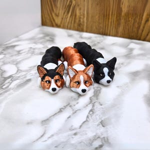 May include: Three 3D printed figurines of corgis in different colors: black and white, brown and white, and copper and white. The corgis are lying down with their heads resting on the surface.