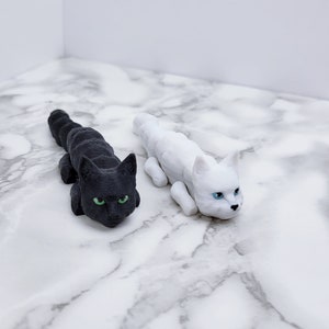 Op de afbeelding: Twee kleine kattenbeeldjes, een zwarte met groene ogen en een witte met blauwe ogen, liggend op een wit marmeren oppervlak.