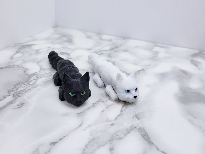 Op de afbeelding: Twee kleine kattenbeeldjes, een zwarte met groene ogen en een witte met blauwe ogen, liggend op een wit marmeren oppervlak.