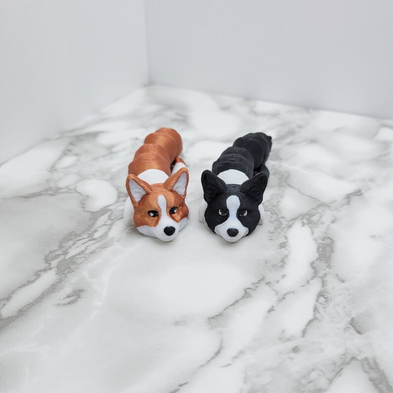May include: Two small, 3D printed, corgi dog figurines. One is brown and white, the other is black and white. Both are lying on a white marble surface.