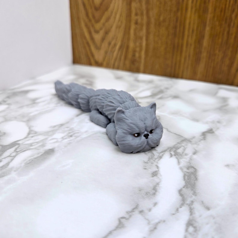 Op de afbeelding: Een grijs, keramiek beeldje van een kat die op zijn zij ligt met zijn kop op de grond. De kat heeft een plat gezicht en grote, ronde ogen.