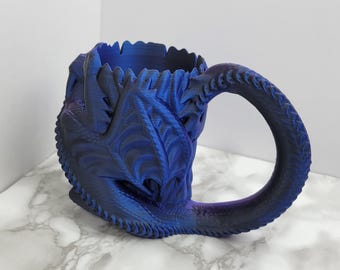 May include: A blue ceramic mug shaped like a dragon's head with a detailed, textured design. The mug has a handle that is shaped like a dragon's tail.