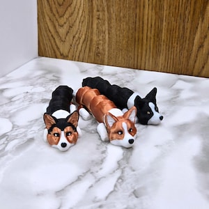 May include: Three 3D printed figurines of corgis in different colors. The corgis are lying down with their heads turned to the side. The corgis are brown and white, black and white, and orange and white.