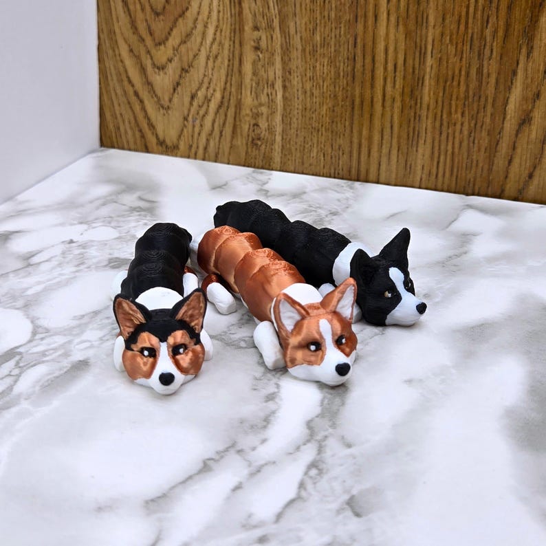 May include: Three 3D printed figurines of corgis in different colors. The corgis are lying down with their heads turned to the side. The corgis are brown and white, black and white, and orange and white.