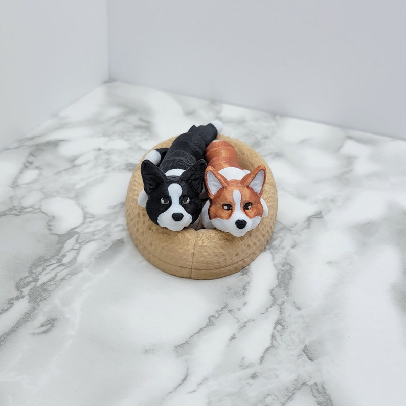 May include: Two plush corgi dogs, one black and white and one brown and white, are lying in a tan-colored dog bed.