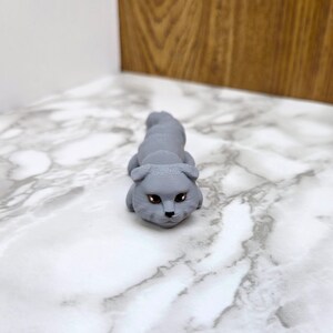 Op de afbeelding: Een grijs 3D-geprint beeldje van een kat met bruine ogen. De kat ligt op zijn buik met zijn kop omhoog en kijkt naar de camera.