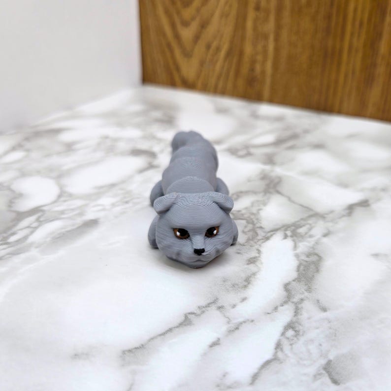 Op de afbeelding: Een grijs 3D-geprint beeldje van een kat met bruine ogen. De kat ligt op zijn buik met zijn kop omhoog en kijkt naar de camera.