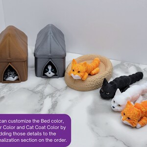 Op de afbeelding: Een set van drie kattenmanden, een bruin, een grijs en een beige. De manden zijn gevormd als kleine huisjes met een kat die uit de ingang kijkt. Er liggen ook vier katten, een oranje, een zwarte, een witte en een oranje-witte. De tekst op de afbeelding luidt: "U kunt de kleur van het bed, de kleur van de drager en de kleur van de vacht van de kat aanpassen door deze details toe te voegen aan het personalisatiesectie van de bestelling."