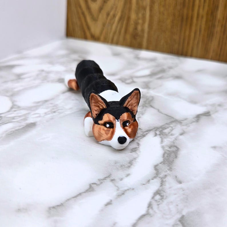 May include: A 3D printed figurine of a black and white corgi dog with brown ears. The dog is lying down with its head resting on the ground.