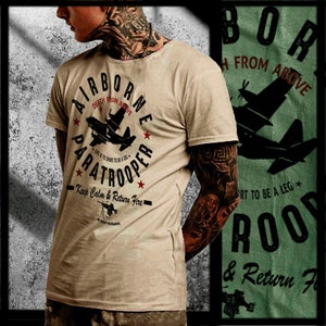 May include: Tan t-shirt with the words "AIRBORNE PARATROOPER" in a circular design, featuring a plane graphic and the phrases "Death From Above" and "Keep Calm & Return Fire." The shirt has a crew neck and short sleeves.