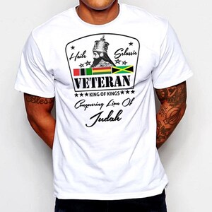 Puede incluir: Camiseta blanca con un gráfico negro y verde que presenta un retrato de Haile Selassie, el texto "Hail Selassie", "VETERAN", "KING OF KINGS", "Conquering Lion Of Judah" y las banderas de Etiopía y Jamaica.