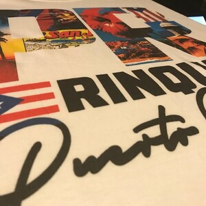 May include: White t-shirt with a black and white graphic design featuring the text "Puerto Rico" and a red, white, and blue Puerto Rican flag. The graphic design also includes a collage of images of Puerto Rico.