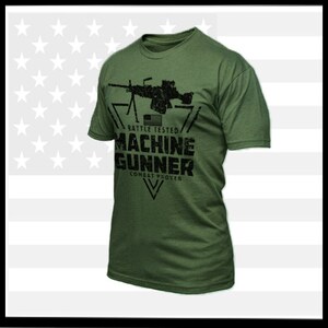 以下が含まれることがあります： マシンガンと「Battle Tested Machine Gunner Combat Proven」というテキストが黒で描かれた緑色のTシャツです。