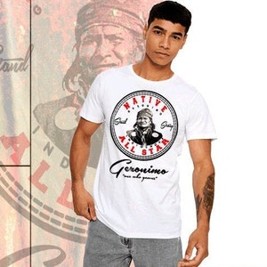 Puede incluir: Camiseta blanca con un gráfico circular rojo y negro que presenta a un hombre nativo americano con el texto "NATIVE", "GERONIMO", "ALL STAR" y "once woke, you're awake".