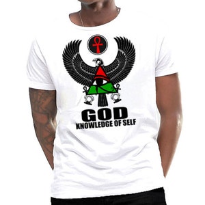 Peut inclure: T-shirt blanc avec un motif noir et vert d'un dieu égyptien avec des ailes, un œil et un symbole ankh. Le texte "GOD KNOWLEDGE OF SELF" est imprimé sous le motif.