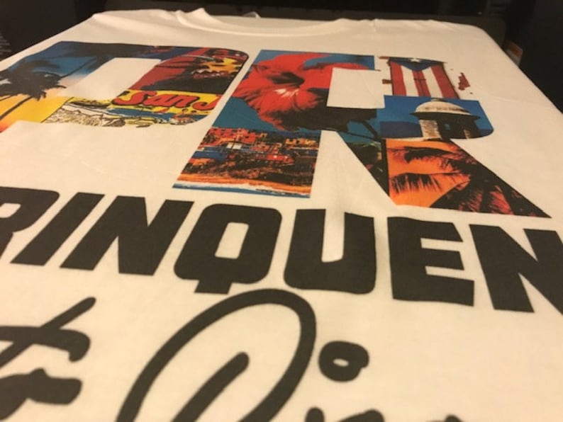 May include: White t-shirt with a graphic design of the letters "DR" made up of images of tropical scenes, palm trees, and the Puerto Rican flag. The text "RINQUEN" is printed in black below the letters.
