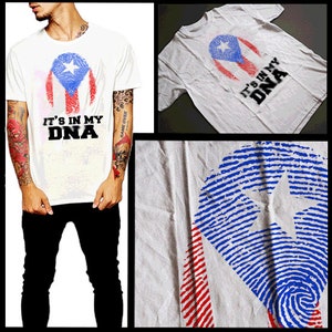 Puede incluir: Camiseta blanca con un diseño de bandera puertorriqueña azul y roja en forma de huella dactilar. El texto "It's in my DNA" está impreso en negro debajo del diseño.