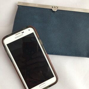 May include: A white Samsung smartphone with a black screen in a clear case is lying on a white surface next to a dark blue wallet with a silver clasp.