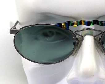 Könnte beinhalten: Ovale Sonnenbrille mit silbernem Metallrahmen und grünen Gläsern. Die Brücke der Sonnenbrille hat ein farbenfrohes, mehrfarbiges Kunststoffdesign.