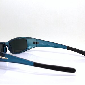 May include: Teal-colored sunglasses with black temples. The sunglasses have a curved design and dark lenses. The text "DUTCH 675" is printed on the frame. The sunglasses are shown against a white background.