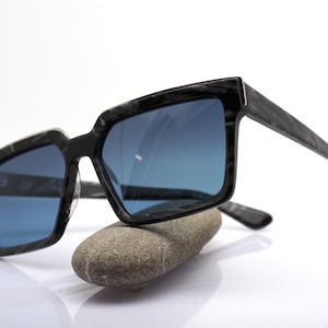 May include: Black sunglasses with a dark blue lens and a black and white marbled frame. The sunglasses are resting on a gray rock.