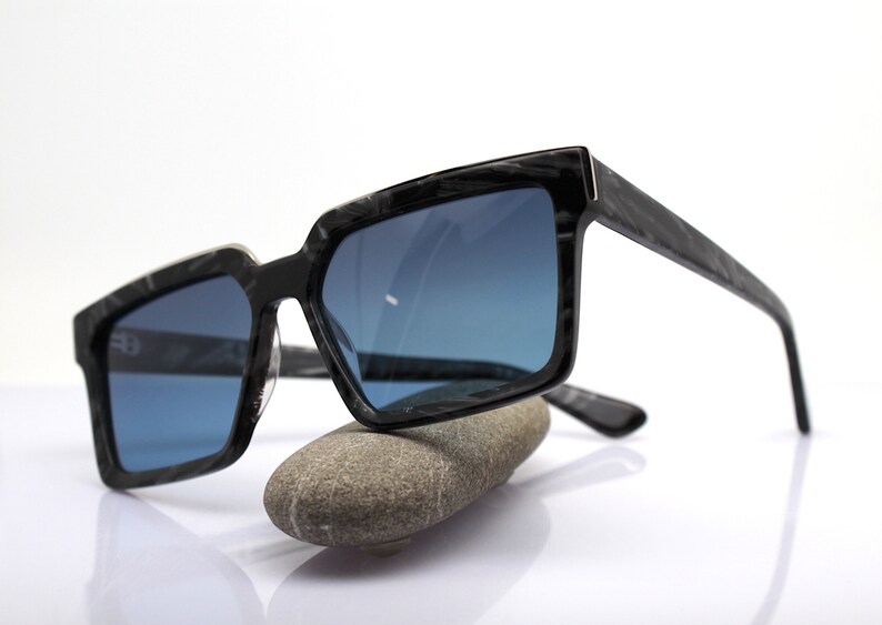 May include: Black sunglasses with a dark blue lens and a black and white marbled frame. The sunglasses are resting on a gray rock.