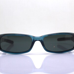 May include: Teal-colored sunglasses with dark lenses. The rectangular frames have a slightly curved design. The sunglasses are set against a white background, with a subtle reflection visible below.