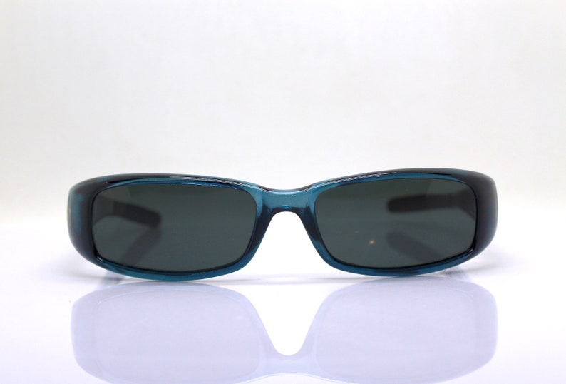 May include: Teal-colored sunglasses with dark lenses. The rectangular frames have a slightly curved design. The sunglasses are set against a white background, with a subtle reflection visible below.