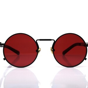 K&ouml;nnte beinhalten: Runde Sonnenbrille mit schwarzem Metallrahmen und leuchtend roten Gl&auml;sern. Die Brille hat ein klassisches Design mit Steg und B&uuml;geln. Die Gl&auml;ser haben eine kr&auml;ftige, einfarbige Farbe und bilden einen auff&auml;lligen Kontrast zum schwarzen Rahmen.