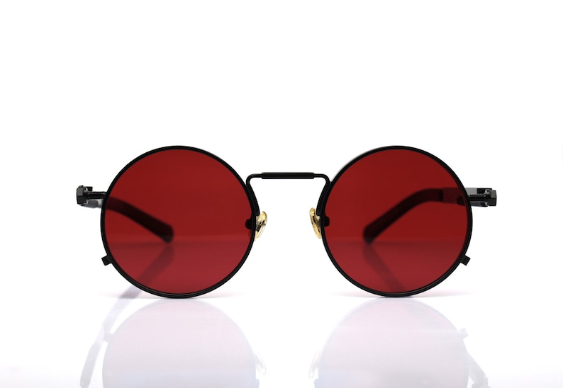 K&ouml;nnte beinhalten: Runde Sonnenbrille mit schwarzem Metallrahmen und leuchtend roten Gl&auml;sern. Die Brille hat ein klassisches Design mit Steg und B&uuml;geln. Die Gl&auml;ser haben eine kr&auml;ftige, einfarbige Farbe und bilden einen auff&auml;lligen Kontrast zum schwarzen Rahmen.
