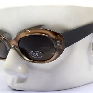 May include: Brown oval sunglasses with dark lenses and a black frame. The sunglasses have a label on the right lens that reads "Optical Protection CE 100% UV Quality".