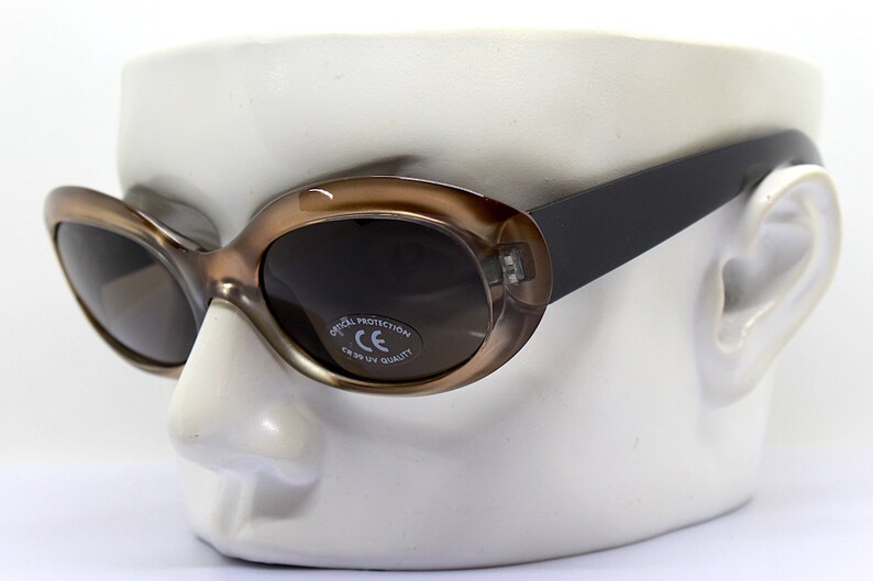 May include: Brown oval sunglasses with dark lenses and a black frame. The sunglasses have a label on the right lens that reads "Optical Protection CE 100% UV Quality".