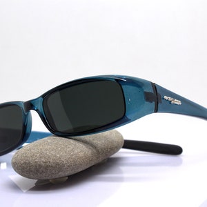 May include: Teal-colored sunglasses with dark lenses. The sunglasses have a curved design and are resting on a gray stone. The temples have a silver accent. The sunglasses are designed to protect the eyes from the sun.