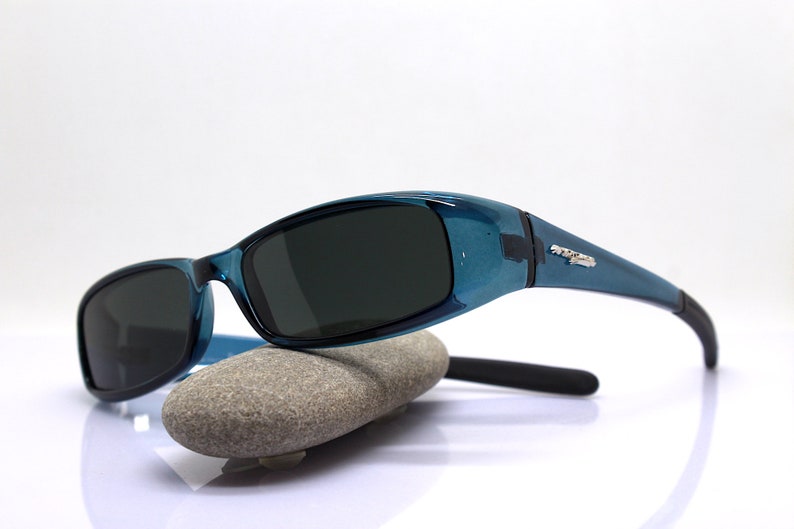 May include: Teal-colored sunglasses with dark lenses. The sunglasses have a curved design and are resting on a gray stone. The temples have a silver accent. The sunglasses are designed to protect the eyes from the sun.