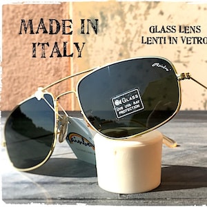 Può includere: Occhiali da sole con montatura in metallo dorato e lenti in vetro verde scuro. Le lenti presentano un adesivo con la scritta GLASS UVB UVA RAY PROTECTION e il marchio Rainbow. Il testo sull'immagine riporta MADE IN ITALY e GLASS LENS LENTI IN VETRO.
