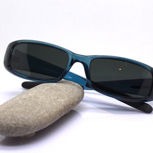 May include: Teal-colored sunglasses with dark lenses rest on a smooth, gray stone. The sunglasses have a rectangular frame and black temples. The stone is oval-shaped and sits on a white background. The image is well-lit.