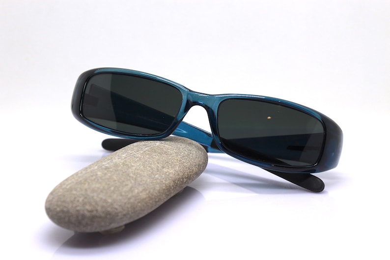 May include: Teal-colored sunglasses with dark lenses rest on a smooth, gray stone. The sunglasses have a rectangular frame and black temples. The stone is oval-shaped and sits on a white background. The image is well-lit.