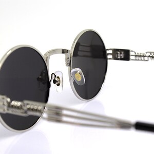 K&ouml;nnte beinhalten: Silberne runde Sonnenbrille mit schwarzen Gl&auml;sern und einem Federscharnier. Die Sonnenbrille hat ein minimalistisches Design und ist aus Metall.