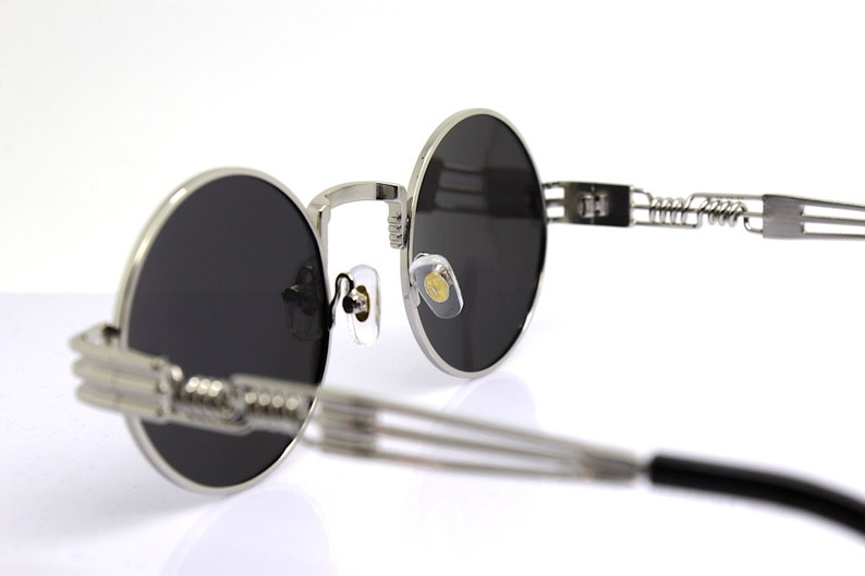 K&ouml;nnte beinhalten: Silberne runde Sonnenbrille mit schwarzen Gl&auml;sern und einem Federscharnier. Die Sonnenbrille hat ein minimalistisches Design und ist aus Metall.