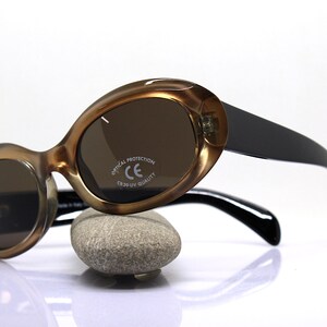 May include: Brown and black oval-shaped sunglasses with brown lenses. The sunglasses have a black plastic frame and black arms. The right lens has the text "OPTICAL PROTECTION CE CR39 UV QUALITY" printed on it.