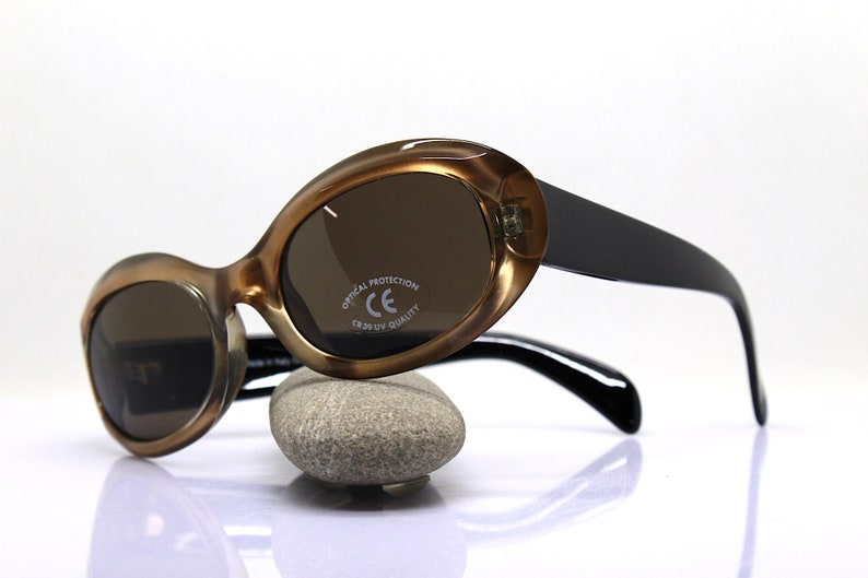 May include: Brown and black oval-shaped sunglasses with brown lenses. The sunglasses have a black plastic frame and black arms. The right lens has the text "OPTICAL PROTECTION CE CR39 UV QUALITY" printed on it.