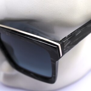 May include: Black square sunglasses with a white stripe on the frame. The sunglasses have a carbon fiber pattern on the arms.