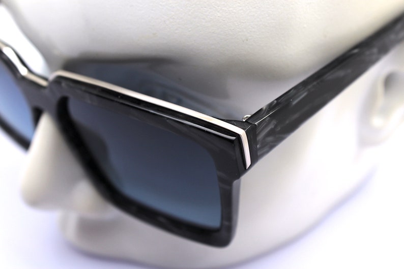 May include: Black square sunglasses with a white stripe on the frame. The sunglasses have a carbon fiber pattern on the arms.