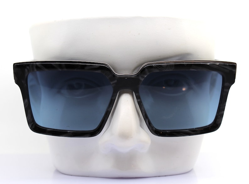May include: Black square-shaped sunglasses with a dark blue lens. The sunglasses are on a white mannequin head.