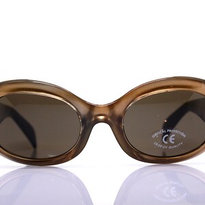 May include: Brown oval-shaped sunglasses with brown lenses. The sunglasses have a white label on the right lens that says "Optical Protection CE  CR39 UV Quality".