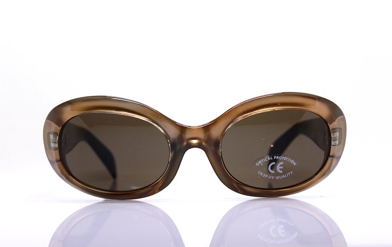 May include: Brown oval-shaped sunglasses with brown lenses. The sunglasses have a white label on the right lens that says "Optical Protection CE  CR39 UV Quality".