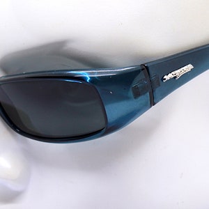 May include: Teal-colored sunglasses with dark lenses. The frames have a glossy finish and a small silver emblem on the temples. The arms are black. The sunglasses are set against a white background.